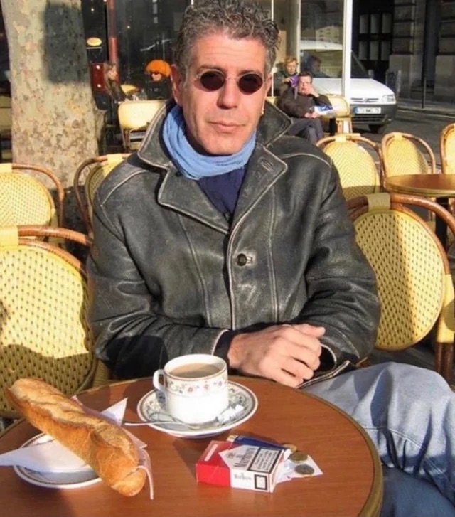 Anthony Bourdain in sunglasses and a leather jacket at an outdoor cafe with coffee, a baguette, and a pack of cigarettes.