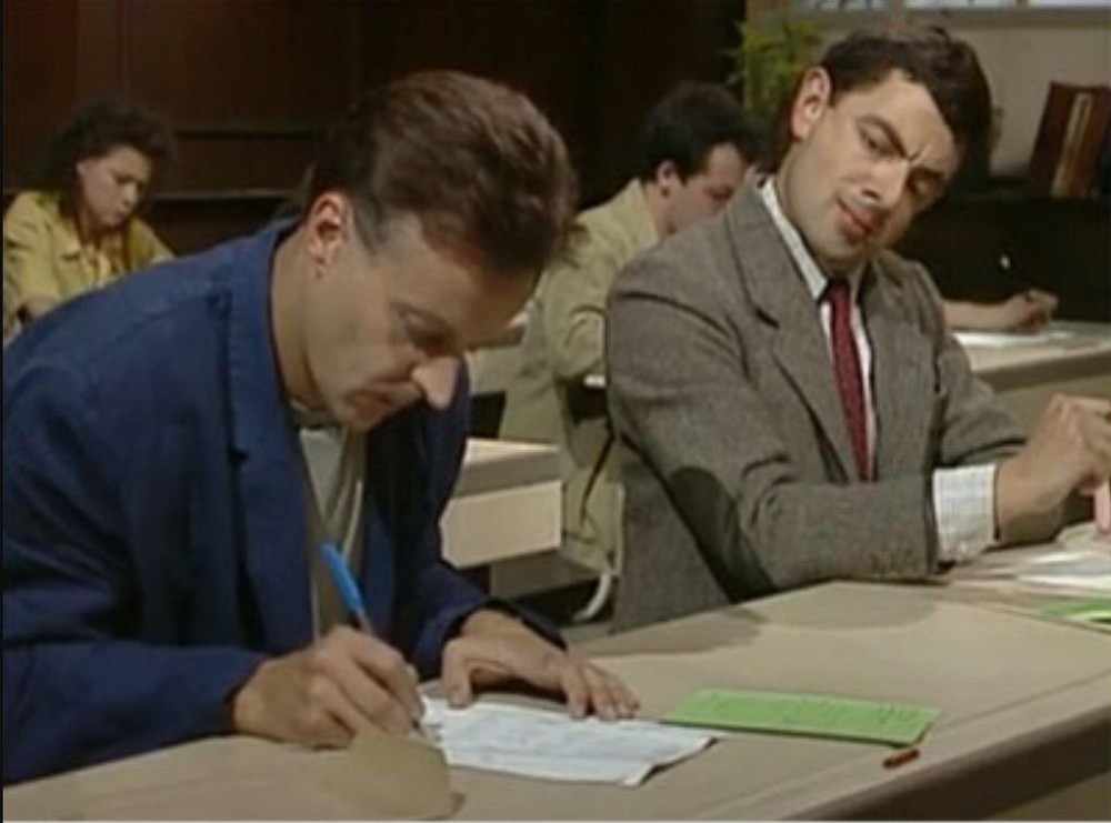 Mr Bean in a grey suit glancing sideways at a man in a blue jacket who is writing an exam.