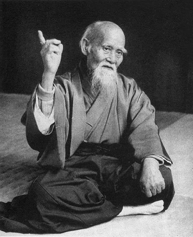Black-and-white photo of Morihei Ueshiba, elderly bearded aikido founder, sitting in seiza and raising one index finger.